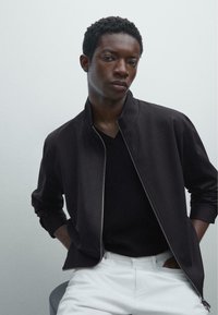 Black zip-up bomber jacket over a black V-neck shirt, with a smooth texture. White pants. Simple design, close-fitting sleeves.