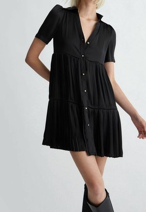 Mujer que lleva un vestido corto negro abotonado con paneles de falda plisada y mangas cortas, combinado con botas negras hasta la rodilla.