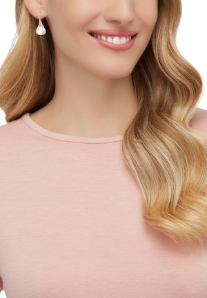 Złote kolczyki wiszące z przezroczystym kryształem w kształcie łzy. Modelka ma luźne, falowane blond włosy i nosi różowy sweter z okrągłym dekoltem.