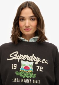 Czarny sweatshirt z logo "Superdry.ca"; posiada kolorowy haft z motywem zachodu słońca i palm, okrągły dekolt i ściągacze przy mankietach.