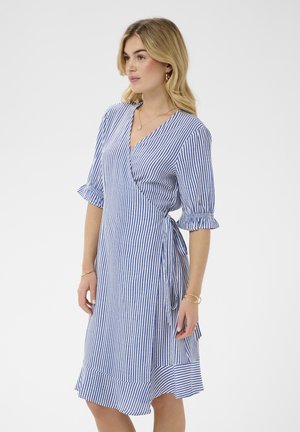 Žena oblečená v modro-bielych vertikálne pruhovaných zavinovacích šatách s volánovými polovičnými rukávmi, bočným zaväzovaním a volánovou sukňou, stojaca na jednoduchom pozadí.