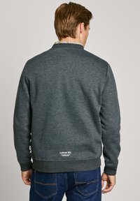 Sudadera gris de corte estándar, con cuello y puños acanalados, y un logo en el dobladillo inferior. Llevada con jeans oscuros y una camisa a cuadros debajo.