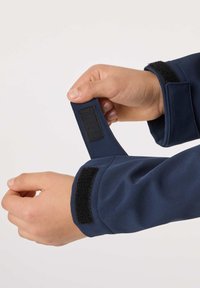 Hands adjusting the navy blue jacket cuff with black Velcro strap against a plain light background.