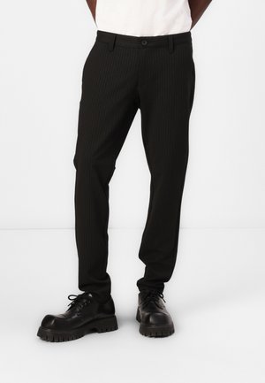 Pantalons noirs à rayures, en tissu, avec une coupe ajustée, des passants de ceinture et une fermeture à bouton. Portés avec des chaussures noires épaisses.