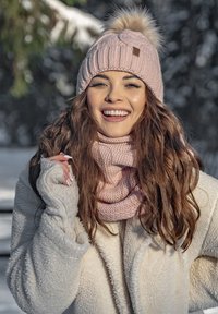 Lichtroze gebreide beanie met een pompon, geribbelde ontwerp en een bruine label. Gecombineerd met een bijpassende gestructureerde sjaal en een crèmekleurige fleece jas.