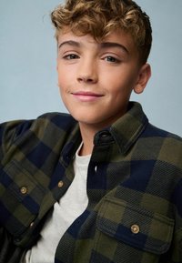 Groene en marineblauwe geruite flanellen overhemd met knoopsluiting, gedragen over een grijze ronde hals t-shirt; krullend haar, neutrale achtergrond.