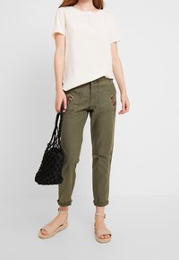 Camiseta de manga corta color crema claro, pantalones verdes olivo con bordado floral y desgastes, bolsa de yute negra, sandalias beige con tiras.