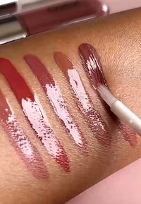 Vier glanzende lipstickstalen in mauve en rozetinten op een arm, waarbij een applicatorborstel de donkerste kleur aanbrengt.