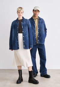 Denim yttertøy med quiltet mønster, blå farge, og trykknapper; sammen med en ribbet kremfarget skjørt og svarte kraftige ankelsko.