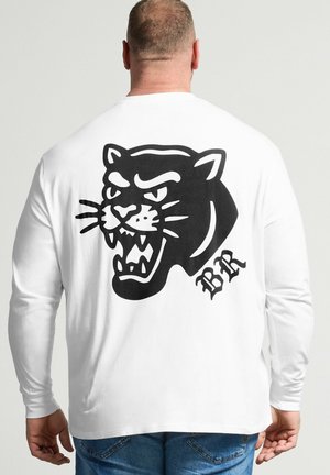 Mężczyzna noszący białą koszulę z długim rękawem z czarną grafiką wściekłego pantery i literami "BR" na plecach, w połączeniu z niebieskimi dżinsami.