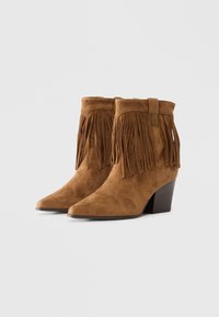 Bocanci din suede maro la glezna, cu franjuri pe partea superioară, vârf ascuțit și toc pătrat. Materialul texturat adaugă profunzime designului.