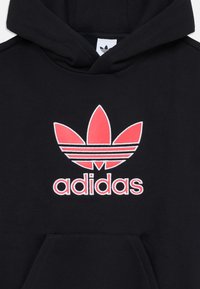 Melna kapuce ar sarkano un baltā Adidas trefoila logo un tekstu. Iezīmējas ar kengūra kabatu un auklu kapucei. Mīksta kokvilnas auduma.