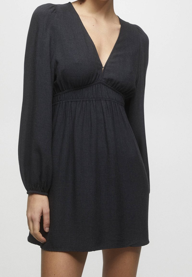 Femme portant une mini robe noire à manches longues avec un décolleté en V et une taille froncée, debout devant un fond clair uni.