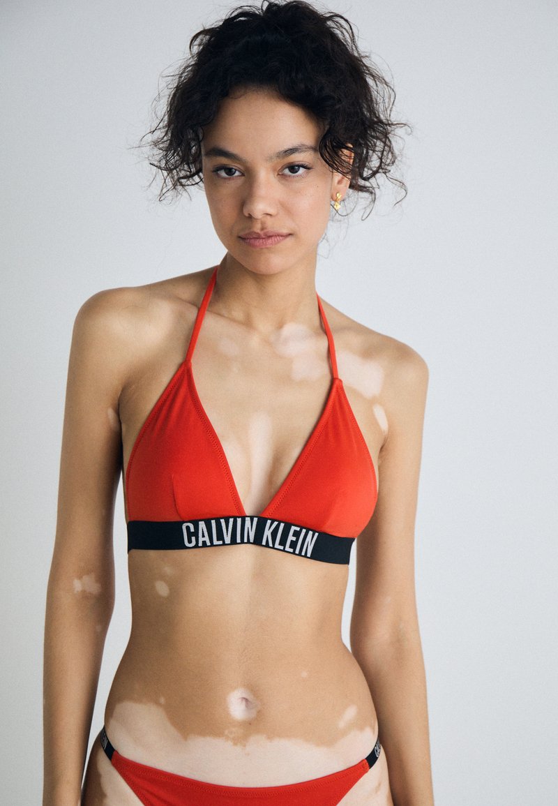 Červený bikiny top s trojuholníkovými pohárikmi, čierny logový pás so nápisom "CALVIN KLEIN." Zodpovedajúce bikiny spodky. Hladká tkanina, minimálne detaily švov.