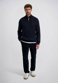 Marineblauer Strickpullover mit Reißverschlusskragen, der ein strukturiertes Fischgrätenmuster aufweist; kombiniert mit dunklen Cordhosen und weißen Sneakers.