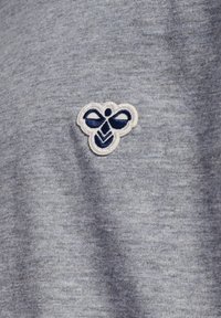 Klein geborduurd donkerblauw en wit bijenembleem gecentreerd op lichtgrijze gebreide stof.