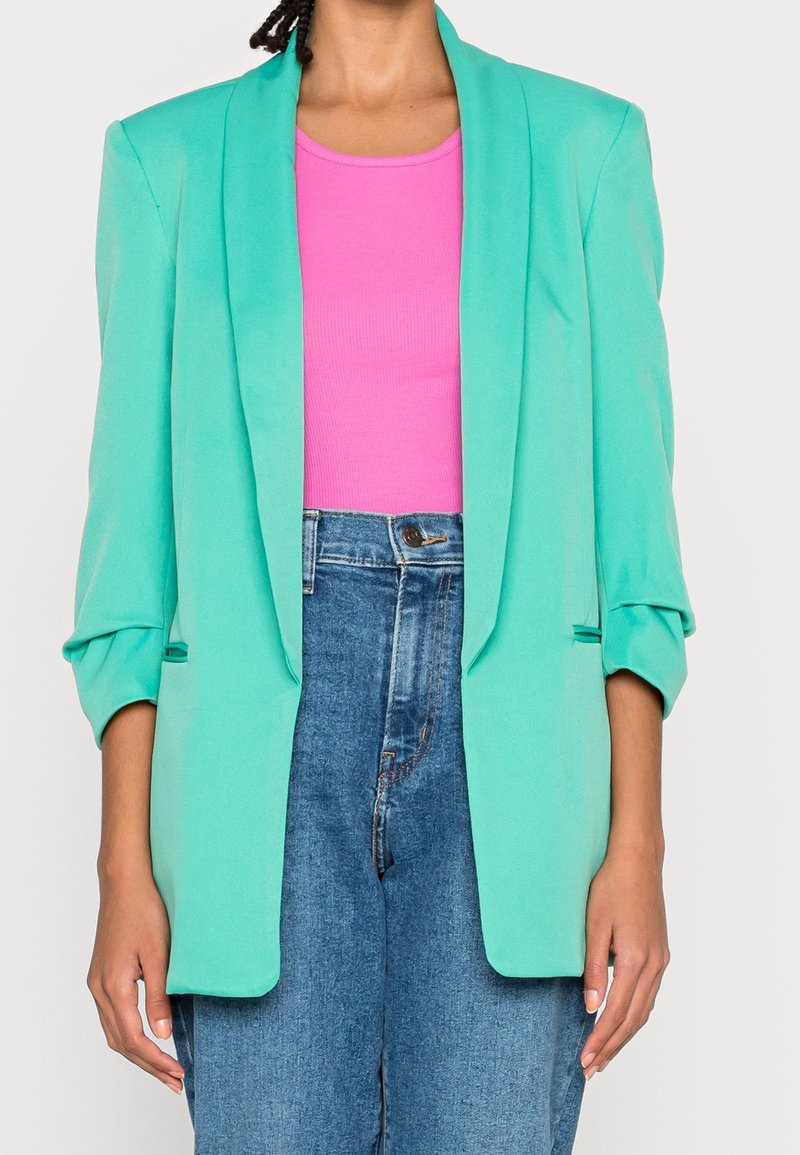 Personne portant un blazer turquoise avec des manches retroussées, un haut sans manches rose vif et un jean bleu taille haute.