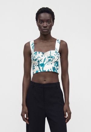 Žena nosiaca biely crop top s modrým kvetinovým vzorom a širokými ramienkami, v kombinácii s čiernymi nohavicami s vysokým pásom, stojaca pred jednoduchým pozadím.