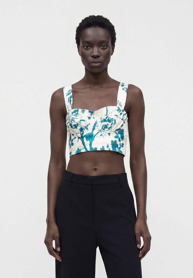 Femme portant un crop top blanc à imprimé floral bleu avec des bretelles larges, associé à un pantalon noir taille haute, debout devant un fond uni.