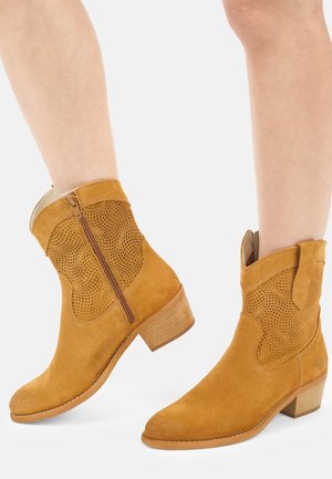 Persoon draagt mid-kuit hoge, lichtbruine suède laarzen met ritssluiting aan de zijkant, decoratief geperforeerd patroon en lage houten blokhakken op een witte achtergrond.