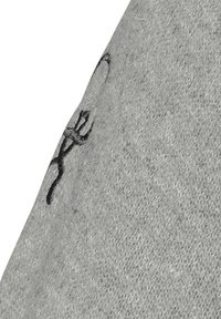 Grå stof med en blød, tekstureret overflade. Har et sort broderet design i øverste højre hjørne, der skildrer en stiliseret figur.