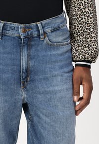 Lyseblå denimjeans med høy midje, fem lommer og bronsefarge detaljer. Har synlig søm og en subtil falming.