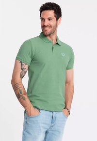 Camisa polo verde con cuello clásico, mangas cortas y un logotipo sutil en el pecho; combinada con jeans de mezclilla azul claro.