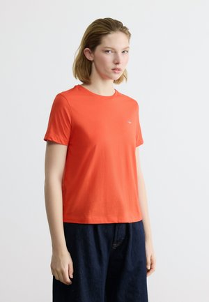 Νεαρό άτομο που φοράει απλό φωτεινό πορτοκαλί κοντομάνικο μπλουζάκι και σκούρο μπλε παντελόνι, στέκεται μπροστά σε απλό φωτεινό φόντο.