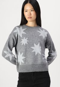 Pulover tricotat gri cu modele de stele gri deschis, decolteu rotund și margine ribbed. Purtați cu pantaloni negri. Textură moale și croială lejeră.