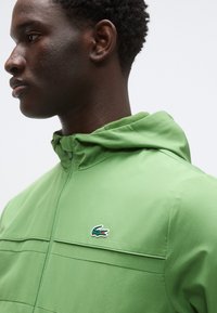 Zelena jopa z zadrgo, ki ima kapuco, teksturirano tkanino in vodoravne šive. Na prsih je opazen značilni logotip Lacoste.