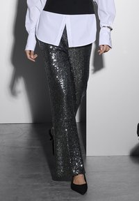 Persoon die loopt in zwarte paillettenbroek, zwarte puntige schoenen en een wit shirt met een zwart vest eroverheen.