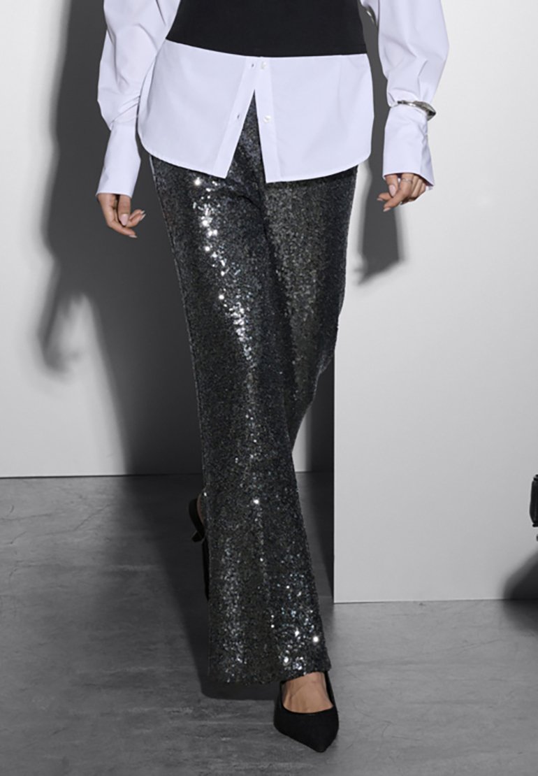 Persoon die loopt in zwarte paillettenbroek, zwarte puntige schoenen en een wit shirt met een zwart vest eroverheen.