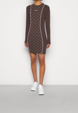 Persoon draagt een nauwsluitende bruine jurk met roze afwerking en geometrisch patroon, gecombineerd met witte sneakers, staand tegen een effen achtergrond.