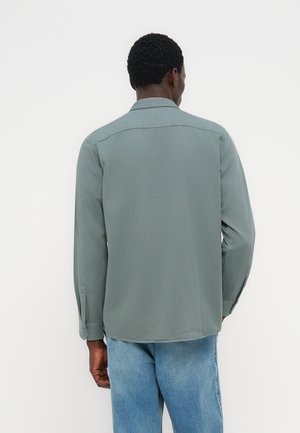 Mann trägt ein langärmliges, salbeigrünes Hemd und hellblaue Jeans, steht mit dem Rücken zur Kamera vor einem schlichten weißen Hintergrund.