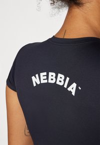 Juodas prigludęs trumpomis rankovėmis viršus su kreive balta "NEBBIA" logotipu ant nugaros. Lygus tekstūros audinys ir apvalus iškirpimas, tinkamas aktyviam sportui.