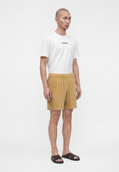 Homme debout portant un t-shirt blanc à manches courtes avec logo, un short texturé couleur sable et des sandales slides marron foncé sur fond blanc.