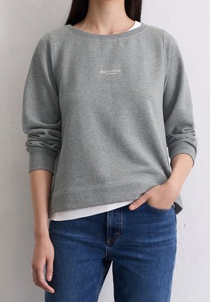 Vrouw draagt een grijze Marc O’Polo trui met lange mouwen over een wit overhemd en blauwe spijkerbroek, met één hand in haar zak.