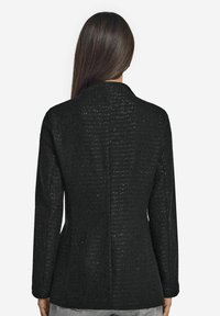 Svart texturerad blazer med en figursydd design, enkel ryggslits och långa ärmar. Tyget har ett vävt mönster och en subtil lyster.