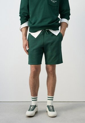 Hombre vistiendo sudadera verde y pantalones cortos a juego, camiseta blanca debajo, calcetines blancos con rayas verdes y zapatillas blancas con detalles verdes.