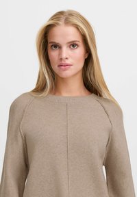 Pull en tricot beige avec un col rond, des manches raglan et des coutures discrètes. Texture douce et design simple pour un look polyvalent.