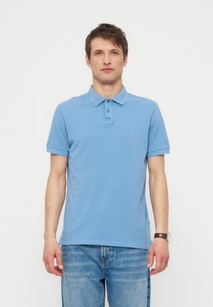 Mężczyzna ubrany w jasnoniebieską koszulkę polo z krótkim rękawem i niebieskie dżinsy, stoi z ramionami swobodnie opuszczonymi na jednolitym białym tle.