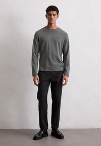 Graues Langarmshirt aus weichem Stoff, mit Rundhalsausschnitt und dezentem Logo-Akzent auf der Brust, kombiniert mit schwarzen schmal geschnittenen Hosen.