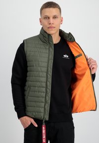 Grön vadderad väst med hög krage, med orange innertyg och två sidofickor. Bärs över en svart sweatshirt.