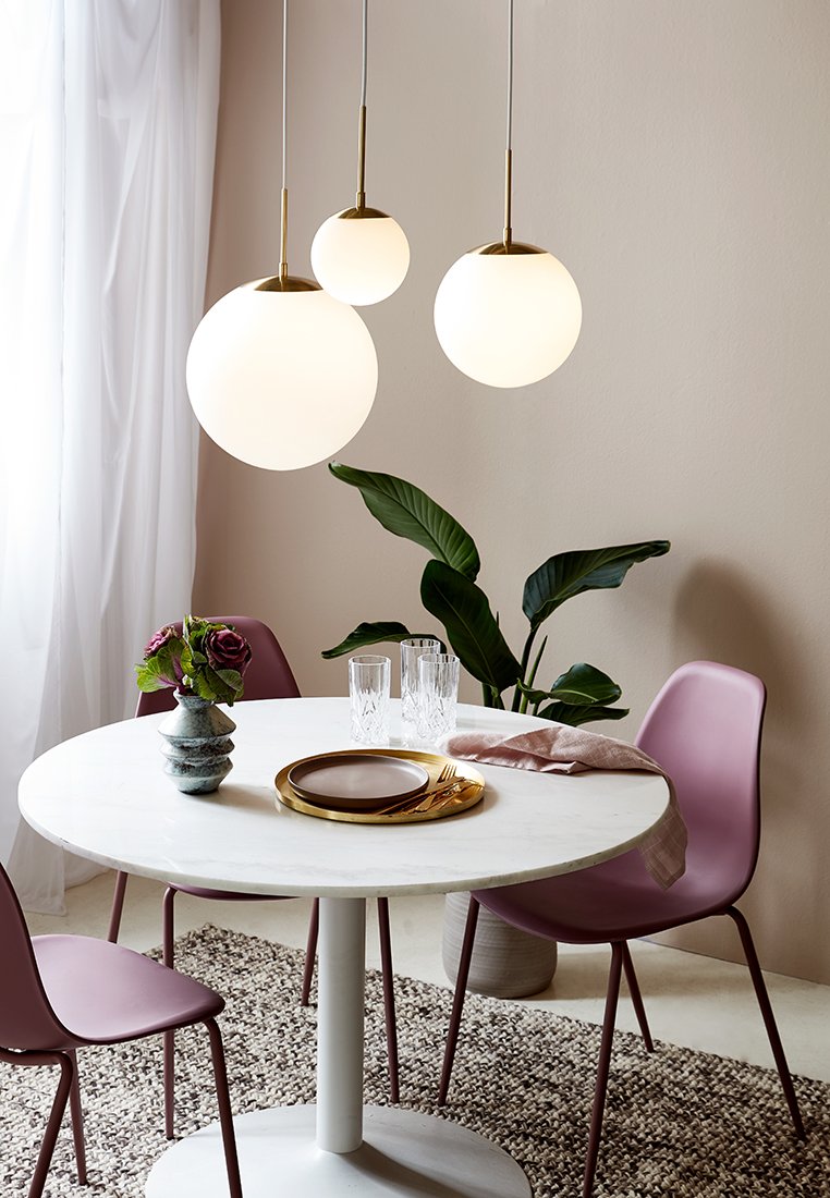 Trzy wiszące lampy w kształcie kul z białymi szkłami i złotymi akcentami nad okrągłym stołem z białego marmuru. W pobliżu różowe krzesła i zielona roślina.