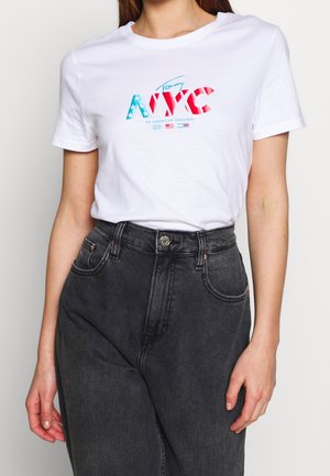 Vrouw draagt een wit T-shirt met de tekst "Tommy NYC" in rood, wit en blauwe sterren en strepen, gecombineerd met zwart, hoog getailleerd spijkerbroek.