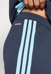 Χέρι που τραβά ένα ναυτικό μπλε αθλητικό ένδυμα, που δείχνει ανοιχτό μπλε τριπλές ρίγες και έναν φερμουάρ τσέπης στο πλάι.