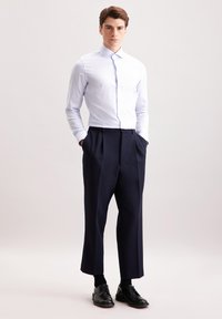 Jasnoniebieska, w białe paski, długa koszula z długim rękawem zestawiona z granatowymi spodniami plisowanymi. Model stoi z rękami w kieszeniach, nosząc czarne buty.