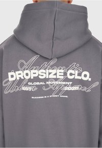 Grauer Hoodie mit weißem, gestapeltem Text auf dem Rücken: "DROPSIZE CLO, Globale Bewegung, Gegründet 2020, Erfolg ist ein Straßenspiel."