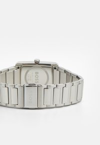 Relógio em aço inoxidável prateado com mostrador retangular, apresentando um fundo liso e um design de bracelete ligado, exibindo a marca "BOSS".