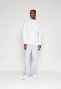 Camicia bianca con bottoni, maniche lunghe, taschino sul petto. Abbinata a jeans blu chiaro e sneakers bianche, su uno sfondo neutro.